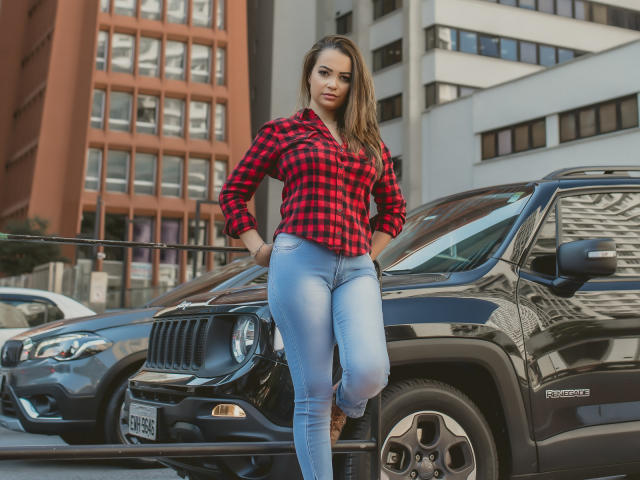 AmaraLove stands with one leg up on a black Jeep, wearing a red plaid shirt and tight blue jeans, showing her ass and a confident look.
