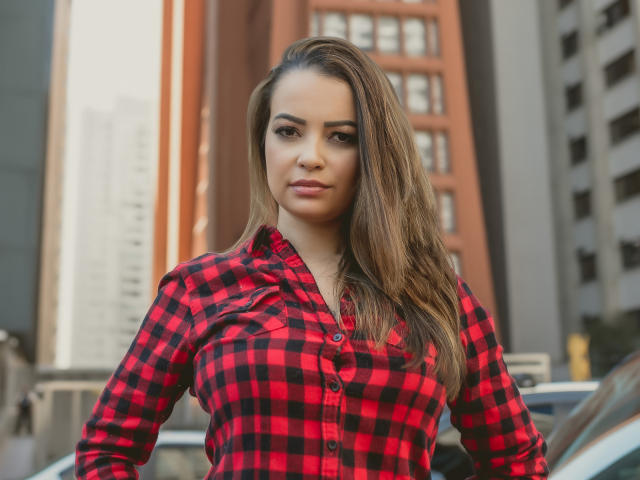 Amara, com longos cabelos castanhos, vestindo uma camisa xadrez vermelha e preta, em um ambiente urbano, com uma expressão confiante e um leve sorriso, exibindo seus seios de forma discreta..
