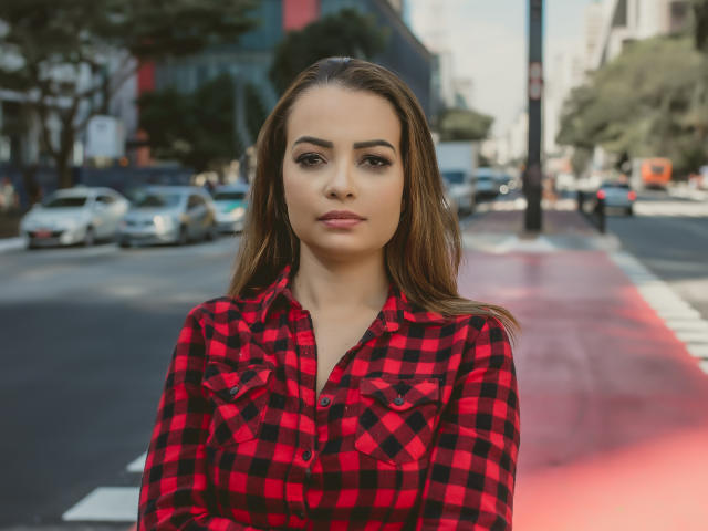Amara, avec ses cheveux bruns, portant une chemise à carreaux rouge et noir, a un visage sérieux et les bras croisés, et se tient sur une rue de la ville, avec des voitures en arrière-plan..