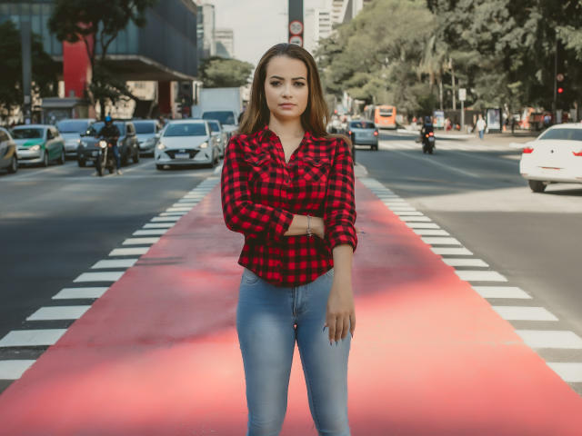 AmaraLove se tient dans une rue de la ville, vêtue d'une chemise à carreaux rouge et noir, d'un jean bleu, et les bras croisés, avec des voitures et des motos en arrière-plan..