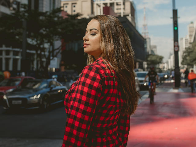 Amara, avec de longs cheveux bruns, une chemise à carreaux rouge et noir, montrant sa poitrine, debout dans une rue de la ville, regardant sur le côté, journée ensoleillée, voitures en arrière-plan..