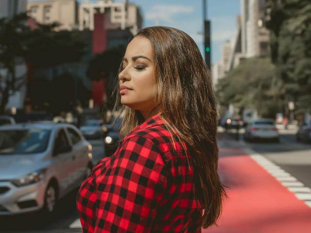 Amara, avec de longs cheveux bruns, portant une chemise à carreaux rouge et noire, se tient sur une rue de la ville, regardant sur le côté avec les yeux fermés, montrant sa poitrine généreuse..