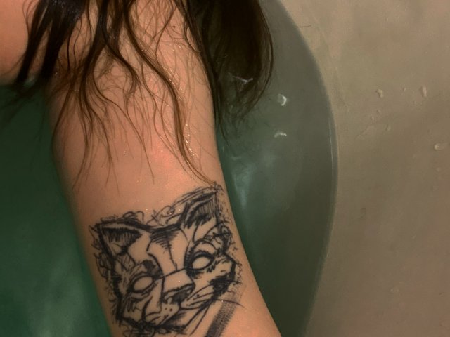 Il braccio di Aleksa Keller immerso in un bagno di colore verde, con goccioline d'acqua sulla pelle, e un tatuaggio a schizzo nero raffigurante il volto di un gatto, con occhi e baffi dettagliati..
