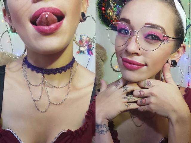 GoddessLufe with pink lipstick, tongue piercing, purple choker, chains, glasses, tattoos, earrings, and rings. She's smiling, hands near face, showing off her pretty pink makeup. Boobs visible.
