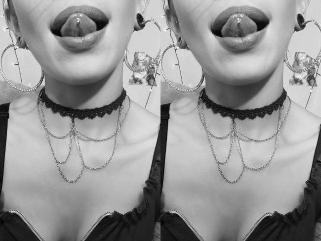 Le visage de GoddessLufe en noir et blanc, la langue tirée, avec un rouge à lèvres épais et foncé, de grandes boucles d'oreilles, un collier en dentelle noire, une chaîne, et un haut noir à volants, laissant entrevoir ses seins..