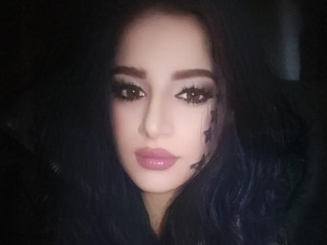 Das Gesicht von KateLaAngel mit glänzenden, rosafarbenen Lippen, dunklem, gewelltem Haar und Smokey-Eye-Make-up, das direkt in die Kamera blickt, mit einem leichten, geheimnisvollen Lächeln..