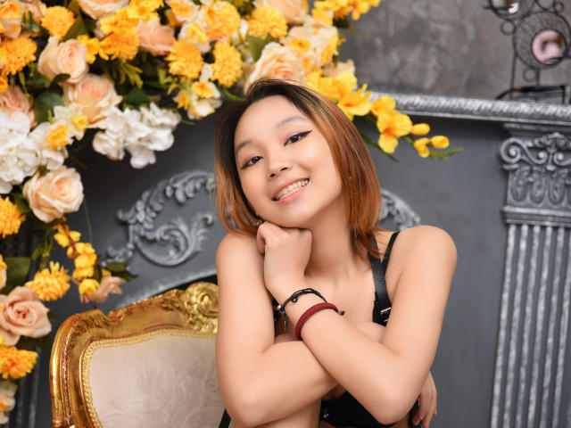 Milla, com cabelo castanho curto, sorrindo, braços cruzados, vestindo uma regata preta, sentada em uma cadeira dourada, cercada por flores amarelas e brancas, com uma aparência fofa e sexy..