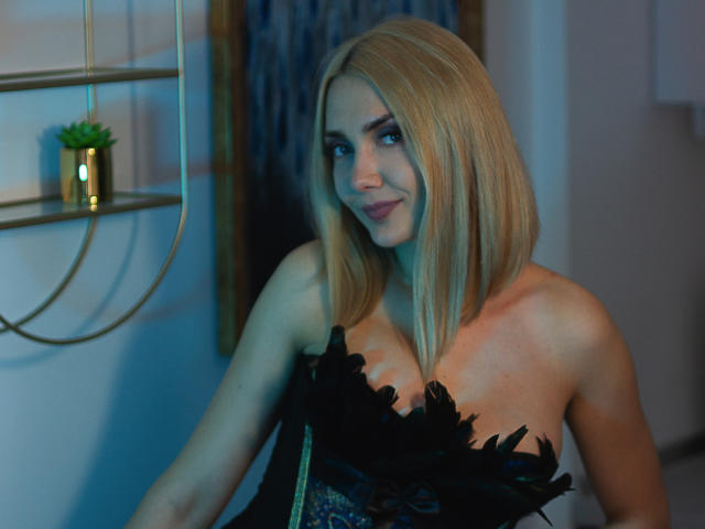 EliseONE has blonde hair, blue eyes, and is wearing a black, strapless dress with flower-like decorations, showing her tits. She's smiling with a slight, sexy, confident look.