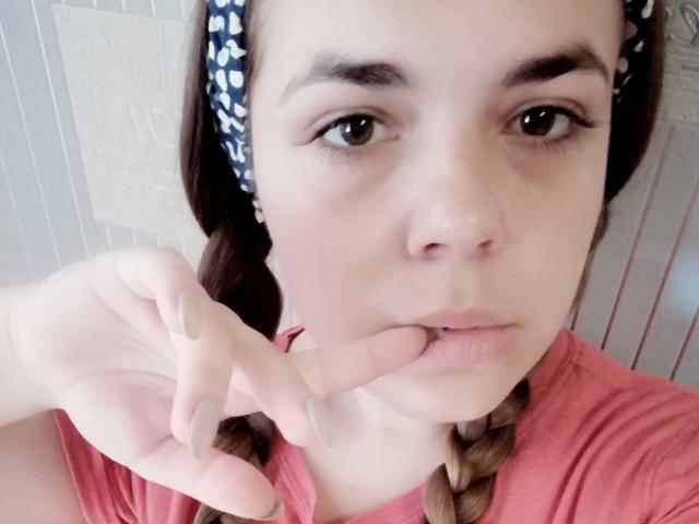 Josefine Darkfeel mit einem nachdenklichen Blick, die an ihrem Finger nagt, trägt ein rosa Hemd, hat geflochtene Haare mit einem blauen, gemusterten Haarreif und beige Nagellack, auf einem Selfie..
