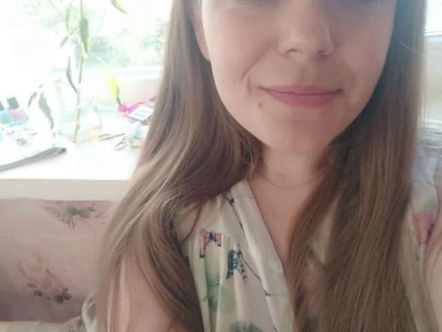 JosefineDarkfeel heeft lichtbruin haar, een subtiele glimlach en een kleine moedervlek onder haar lichtroze lippen. Ze draagt een top met bloemenprint en een dunne ketting..