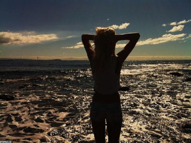 Josefine Darkfeel steht am Strand, mit dem Rücken zur Kamera, die Hände im blonden Haar, sie trägt ein Tanktop und Shorts und ist im Sonnenlicht, das auf den feuchten Sand fällt, nur als Silhouette zu sehen..