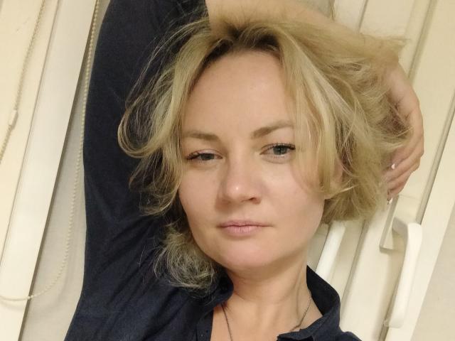 KayaBlonde a les cheveux blonds, légèrement en désordre, elle porte un haut bleu marine, est allongée avec un bras derrière la tête, et a l'air détendue, avec une expression subtile, presque un sourire..