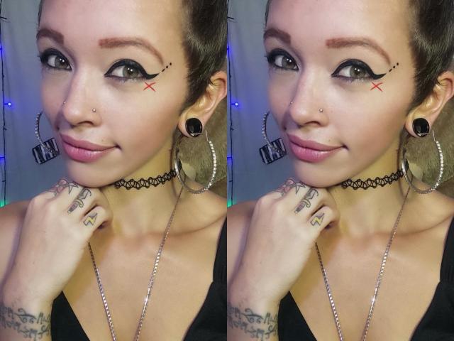 GoddessLufe com delineador preto, tatuagens X perto dos olhos, colar preto, brincos grandes, piercing no nariz e tatuagens nas mãos, sorrindo levemente. Ela tem batom rosa e cabelo castanho..