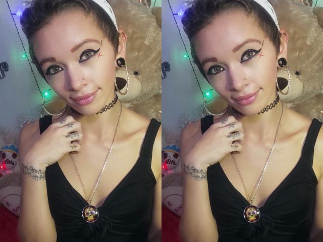 GoddessLufe tem cabelo castanho curto, delineador preto com pontas finas, pequenas tatuagens, colar preto, brincos, vestido preto, colar com uma pedra preciosa, está sorrindo e segurando a mão perto do rosto..