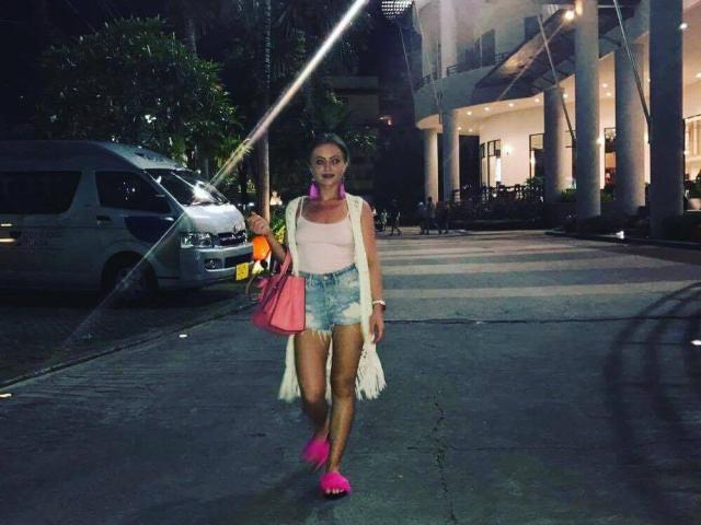 LovelyEvee cammina di notte, indossando una canotta bianca, pantaloncini di jeans sfilacciati, orecchini rosa, tacchi rosa e una borsa rosa, tenendo in mano una bevanda arancione. In secondo piano, si vedono un furgone e un edificio..