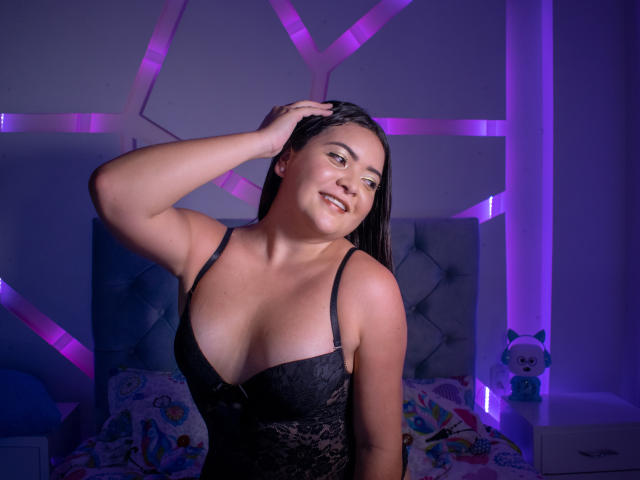 SamanthaWade com seios médios, vestindo uma blusa preta de renda, com a mão no cabelo, sorrindo, luzes LED roxas, cama com travesseiros coloridos, um brinquedo de pelúcia e um rosto fofo..