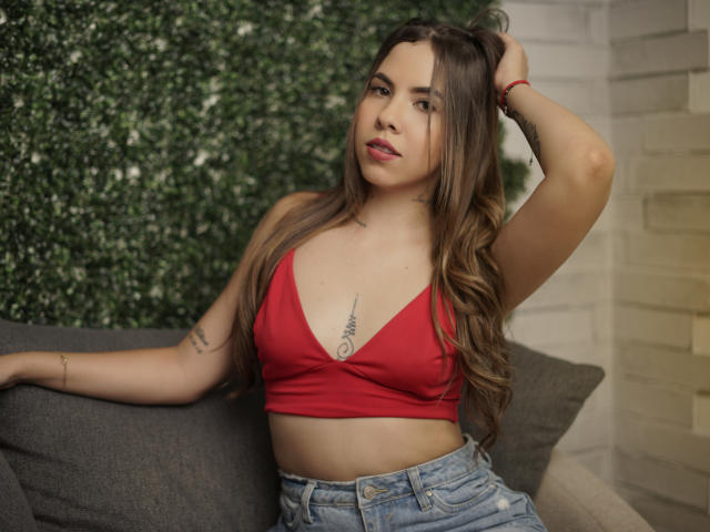Emmy Russo, con lunghi capelli castani, un top rosso e jeans blu, mostra un tatuaggio sotto il seno, con un braccio alzato, seduta su un divano grigio..