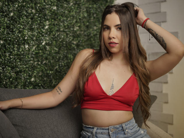 EmmyRusso, con cabello castaño largo, top rojo corto que muestra poco busto, tatuaje en el brazo derecho, pantalones cortos vaqueros, mano en el cabello, fondo de pared verde, sofá gris, pulsera roja..