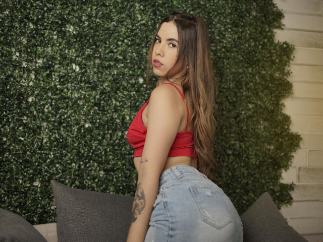 EmmyRusso, con lunghi capelli castani, un top rosso e jeans blu, che mostra il sedere, un tatuaggio sul braccio destro, che guarda oltre la spalla, con una parete verde e cuscini grigi sullo sfondo..