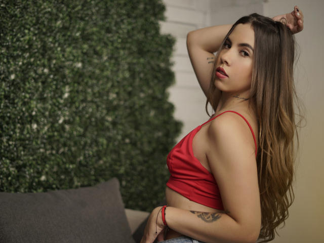 Emmy Russo, avec de longs cheveux bruns, un haut court rouge et un pantalon gris, posant les bras levés, montrant un tatouage sur le côté droit et un bracelet rouge au poignet gauche..