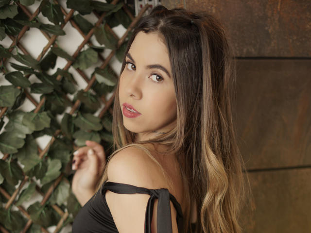 Emmy Russo, con lunghi capelli castani, che si guarda indietro, indossa una maglietta nera con una spalla scoperta, che mette in mostra il seno, ha gli occhi marroni e il rossetto rosa, ed è in piedi di fronte a un traliccio..
