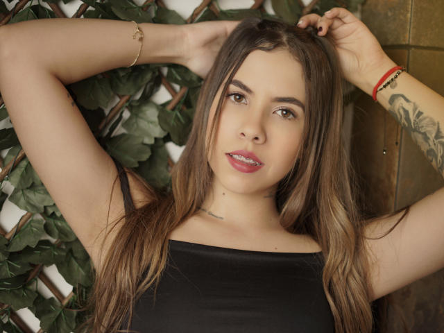 EmmyRusso con le braccia alzate, capelli castani, rossetto rosa, top nero, tatuaggio sull'avambraccio destro, braccialetto dorato, braccialetto rosso, leggero sorriso, occhi castani e una piccola lentiggine sul mento..