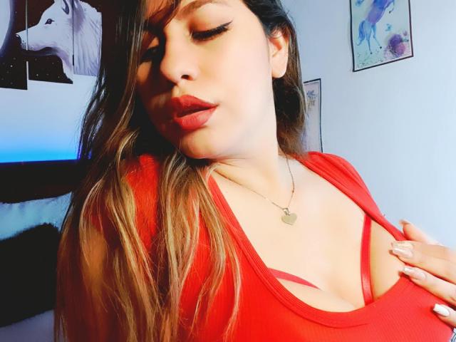 AnastaciaMonaco, com cabelos castanhos longos, batom vermelho e uma blusa vermelha que mostra o colo, com a mão no peito, delineador, colar e uma expressão ligeiramente sedutora..