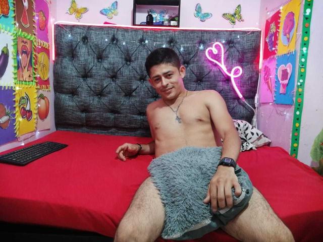 AnthonyHotyBig, sin camisa, con una cara linda, sentado en una cama roja, con una manta gris y suave cubriendo su entrepierna, un corazón neón rosa de fondo, y un reloj negro en la muñeca..