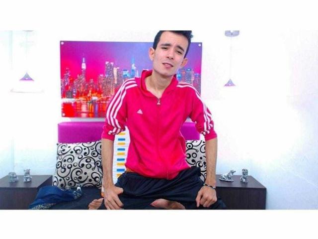 HornyGay69 portant un t-shirt de sport Adidas rouge, assis en tailleur sur un lit avec un pantalon noir, des coussins à motifs noir et blanc, et une affiche colorée représentant un paysage urbain en arrière-plan..