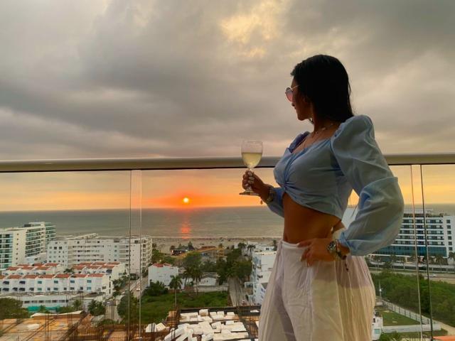 SammyHoffman, avec des seins, portant un débardeur bleu et une jupe blanche, tenant un verre de vin, debout sur un balcon, coucher de soleil sur l'océan, ciel nuageux, ville en contrebas..