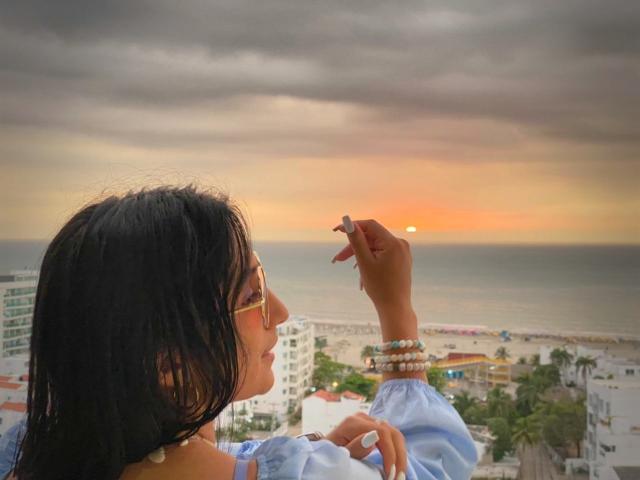 Sammy Hoffman, avec ses cheveux foncés, portant des lunettes et une chemise bleue, tenant une cigarette, regardant un coucher de soleil sur l'océan, portant des bracelets blancs et un collier..
