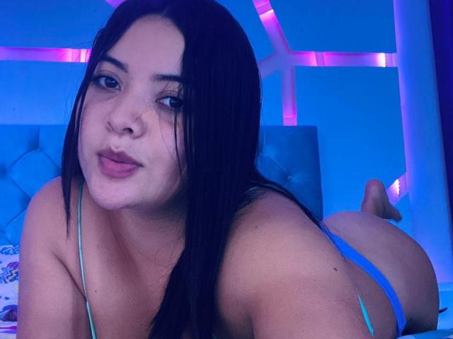 Samantha Wade, con cabello negro largo, labios carnosos y lencería azul, acostada en una cama, mostrando su trasero, mirando a la cámara, en una habitación con iluminación azul..