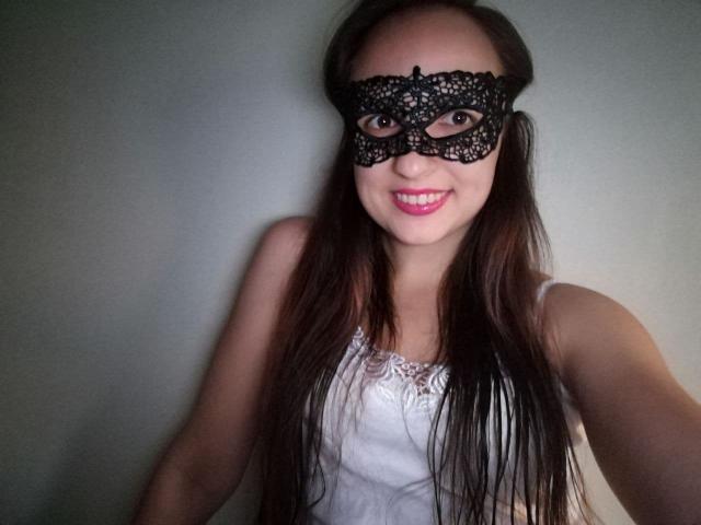 MeganFire69, com cabelos castanhos longos, usando uma máscara preta de renda e uma camiseta branca com detalhes em renda, sorrindo para a câmera com batom rosa. Seios visíveis sob a blusa..