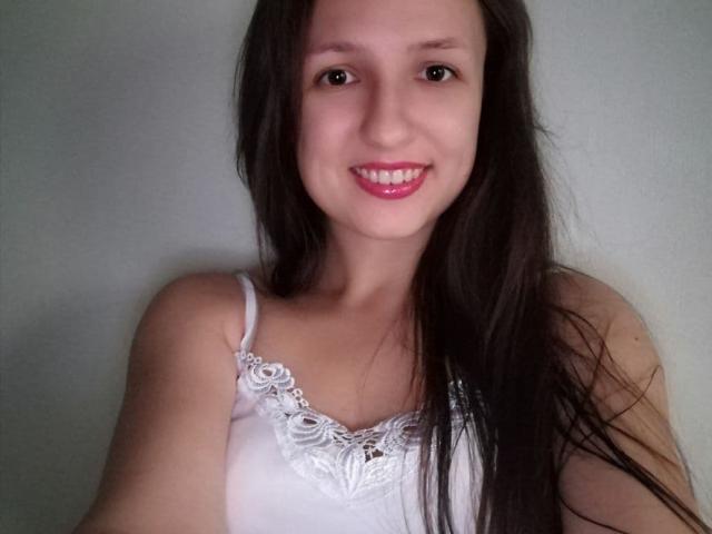 MeganFire69, avec de longs cheveux bruns, un débardeur blanc à dentelle, souriant avec du rouge à lèvres rose, montrant de petites seins, une peau claire et des yeux marrons, dans un autoportrait..