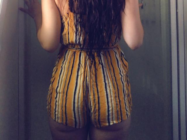Bellefrance69 staat met haar rug naar de camera, ze draagt een geel-zwarte gestreepte jumpsuit die haar achterwerk laat zien, met lang, nat, donker haar, in een slecht verlichte kamer..