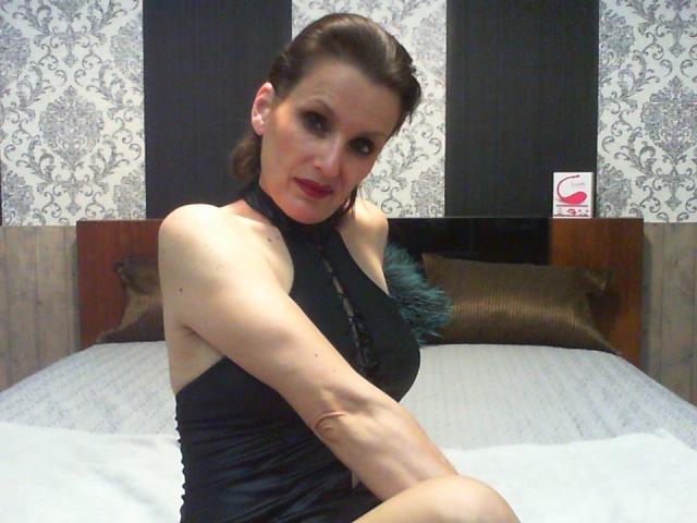 MelindaHottX, wearing a black halter dress, sitting on a bed with her arms crossed, showing her bare shoulders and small tits, with brown hair in a side braid.