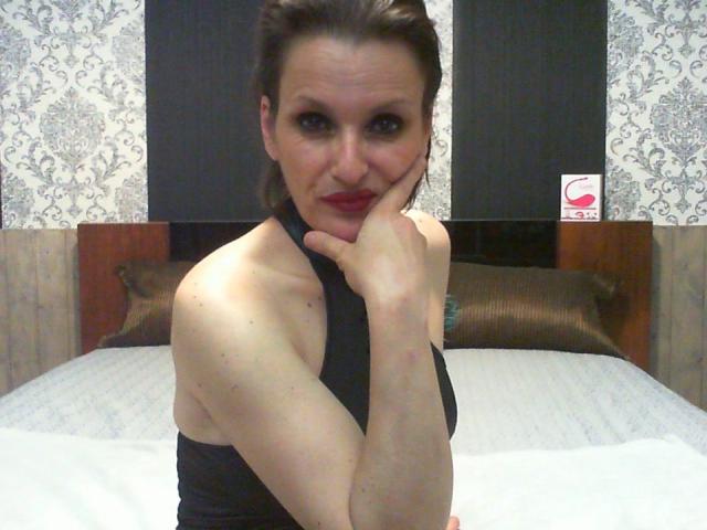 MelindaHottX with dark brown hair, red lipstick, black halter top, showing her bare shoulders and resting her chin on her hand, smiling at the camera. Her tits are partially visible.