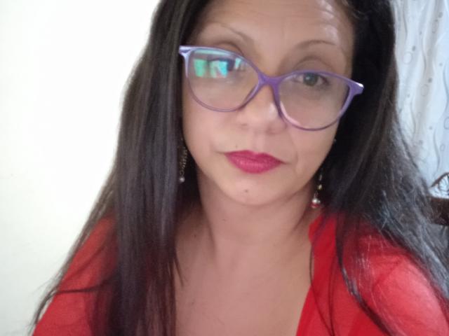 Mistress Sara mit lila Brille, rotem Lippenstift und schwarzem Haar, trägt ein rotes Hemd, zeigt einen kleinen Muttermal auf ihrer rechten Wange und ein leichtes Lächeln..