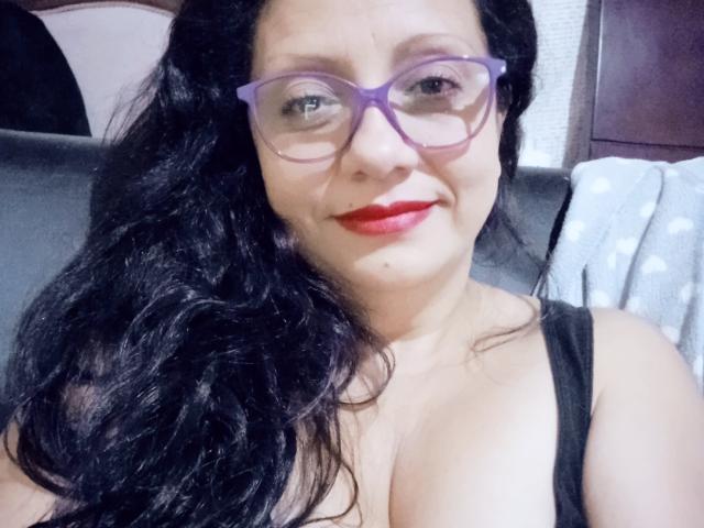 La signorina Sara, con occhiali viola, rossetto rosso e capelli neri ondulati, mostra il suo seno prosperoso in una canotta nera, sorridendo alla fotocamera..