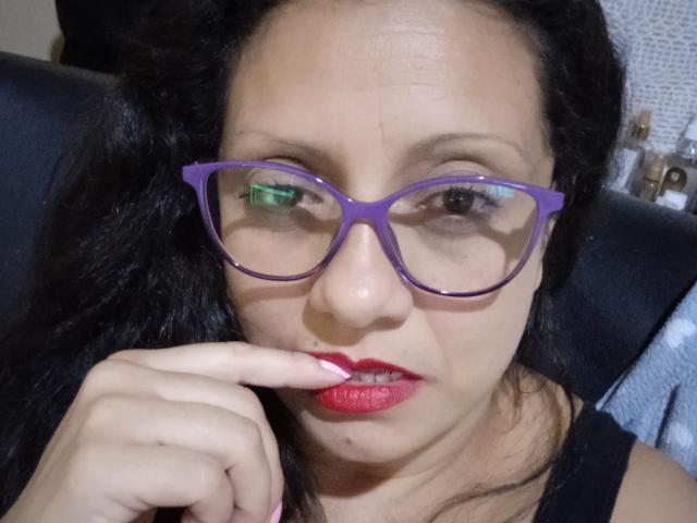 MistresSara with purple glasses, red lipstick, and black hair, biting her finger with pink nails, looking slightly annoyed. Her tits are small, and her skin is tan.