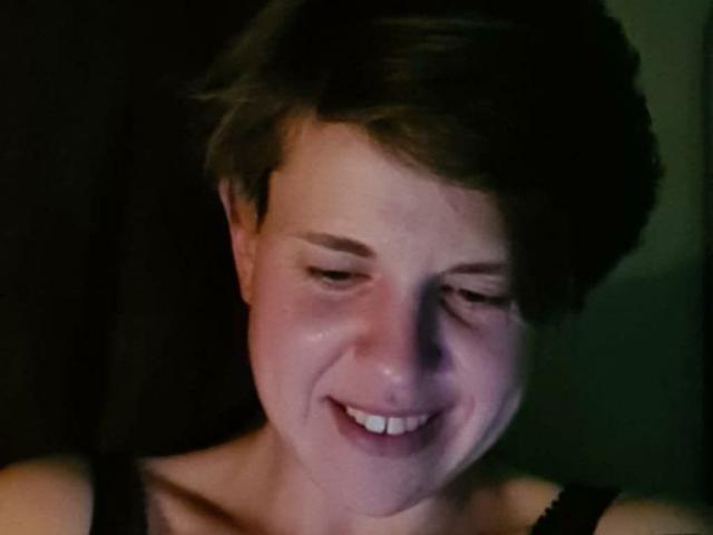 SherlineStone has short brown hair, a smiling face, and visible wrinkles on her forehead. She's wearing a black sleeveless top, and the lighting is dim.