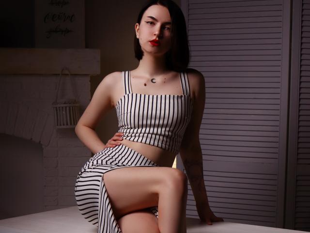 ViolaBlack with pale skin and black hair, wearing striped black and white top and skirt, sitting with one leg up, showing her ass and small tits, red lips, tattoo on chest.