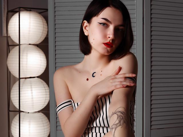 ViolaBlack with short black hair, red lipstick, and striped top, crosses arms, showing tattoos, small moon and dot tattoos on chest, standing in front of soft lanterns.