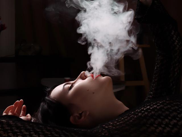 ViolaBlack is smoking, with thick white smoke curling around her head. She's wearing a black lace top, her eyes are closed, and she has dark hair and a small mole.