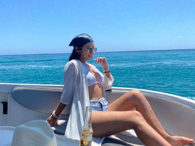 SammyHoffman with tits in white bikini top, denim shorts, and sheer shirt, sitting on boat, legs crossed, holding drink, ocean in background, wearing sunglasses, hat on table.