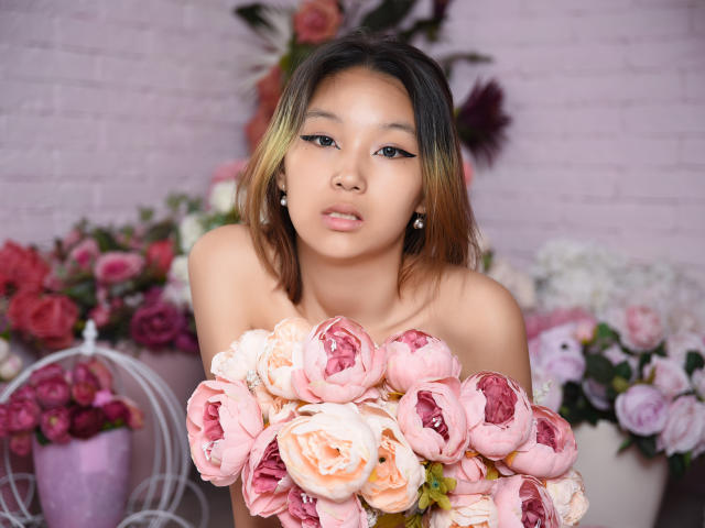 MillaWaves, con un bouquet di rose rosa e pesca, che tiene appoggiato sulle sue spalle scoperte, ha gli occhi marroni e i capelli castano chiaro, con effetto "ombré", indossa orecchini di perle e ha un'espressione neutra..