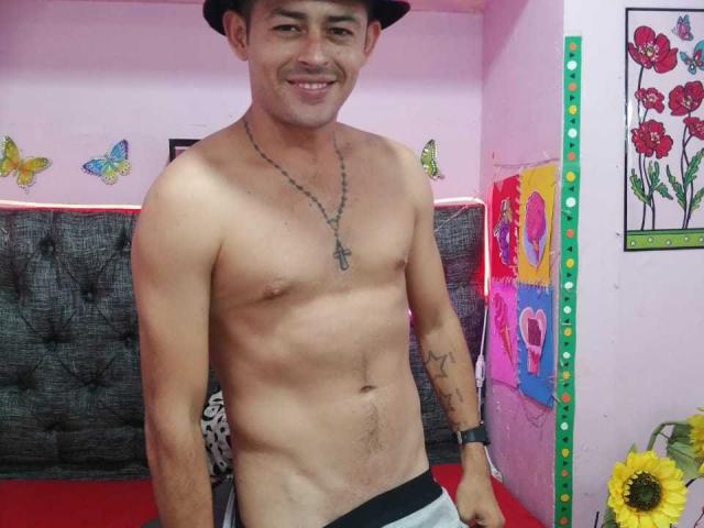 AnthonyHotyBig, sem camisa, com um chapéu preto, colar e tatuagem no pulso, sorrindo, mostrando um peito tonificado, sem seios, peludo, vestindo calças cinzas, em um quarto rosa com decoração de borboletas..
