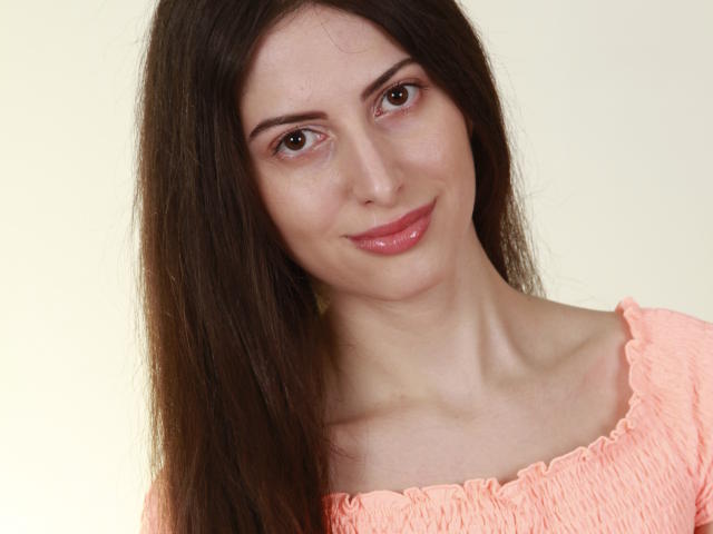 AmaazingLady met lang bruin haar, glanzende roze lippen en een lichte huid, draagt een perzik-oranje top en heeft een schattige glimlach en bruine ogen. Ze kijkt iets naar rechts..