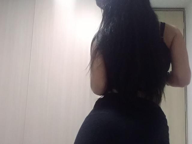 SeleneBrigth está de vuelta, con su largo cabello negro, vistiendo una blusa negra, mostrando sus glúteos con unos pantalones negros ajustados, de pie en una habitación con una pared de madera clara..
