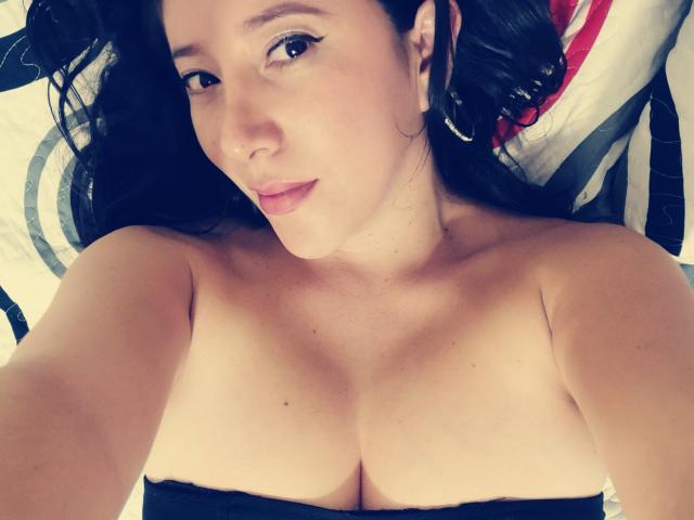 HotPamelaSex with dark curly hair, light skin, and small nose, lying down, topless, showing her bare boobs, subtle makeup, and a slight smile, looking at the camera.